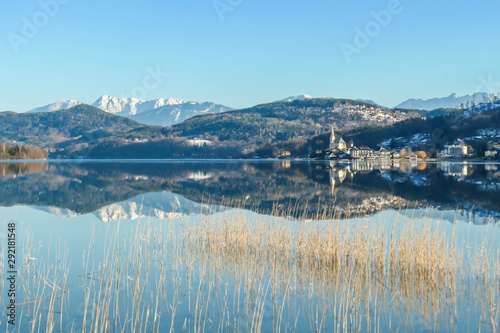A view on a city on the shore of a lake with Alps in the back. View is disturbed by grass. The calm surface of the lake reflects the mountains, sunbeams and cloud. Church with bell tower in the middle