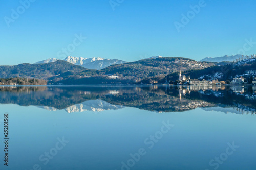 A view on a lake and Alps in the back. The calm surface of the lake is reflecting the mountains  sunbeams and clouds. Little city located at the lake side. Church with bell tower in the middle