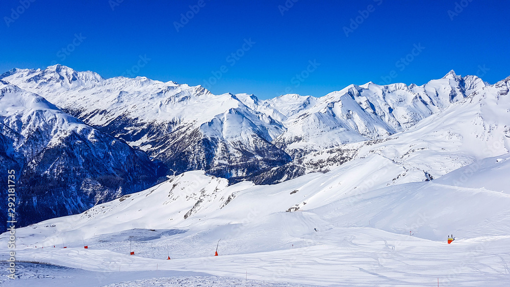A beautiful and serene landscape of mountains covered with snow. Thick snow covers the slopes. Clear weather. Perfectly groomed slopes. Massive ski resort. Red ski poles on the side of the trail.