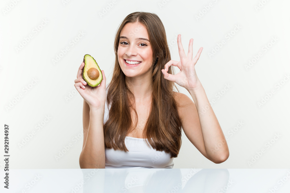 Young caucasian woman holding an avocado cheerful and confident showing ok gesture.