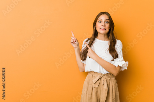 Young caucasian woman shocked pointing with index fingers to a copy space.