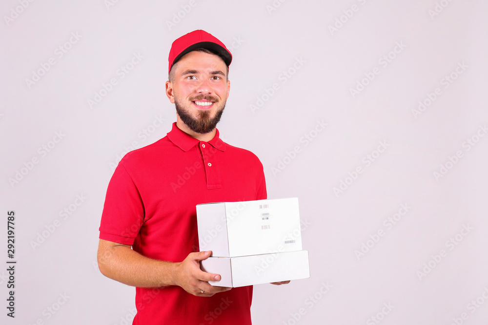Young handsome delivery guy wearing red uniform and cap holding the blank cardboard box over isolated white background. Portrait of friendly bearded man carrying parcel. Copy space for text.