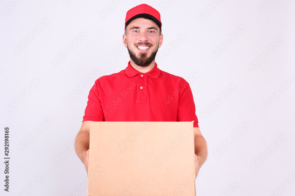 Young handsome delivery guy wearing red uniform and cap holding the blank cardboard box over isolated white background. Portrait of friendly bearded man carrying parcel. Copy space for text. Young handsome delivery guy wearing red uniform and cap holding the blank cardboard box over isolated white background. Portrait of friendly bearded man carrying parcel. Copy space for text.