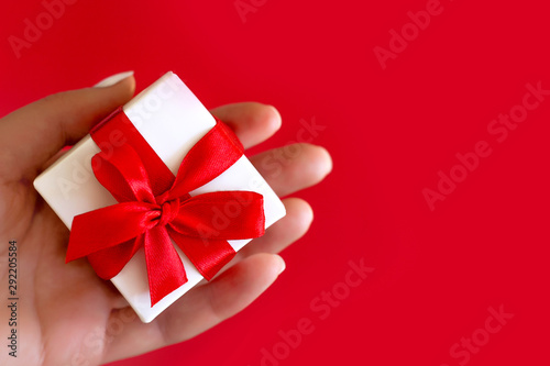 Female hand holds a small white box with a red bow on a red background. Gift in a female hand, copy space
