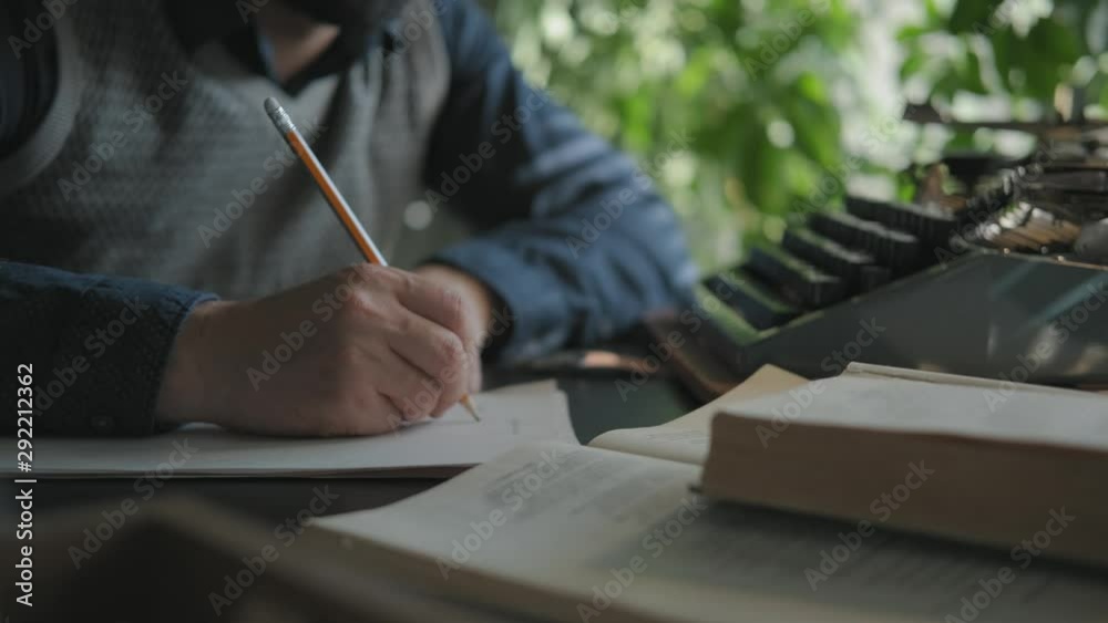 Male writer works sitting at the table. Medium shot: hands, a sheet of paper and a pencil. The writer is working on a draft, writes in pencil. A man writes on paper, typewriter is in front of him