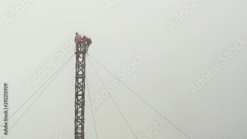 Workers in an orange vest and wearing safety harnesses working on a tower, telephone pole supported by wires and surrounded by fog