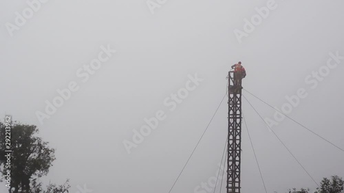 Workers in an orange vest and wearing safety harnesses working on a tower, telephone pole supported by wires and surrounded by fog