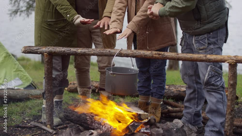Tilt up shot of two senior couples standing by campfire, warming hands above it and talking about something while hiking in forest on autumn day Tilt up shot of two senior couples standing by campfire, warming hands above it and talking about something while hiking in forest on autumn day
