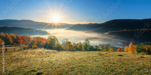 amazing countryside in fall season at sunrise. gorgeous view in to the valley full of fog in morning light. sun above the distant mountain. fence through rural field on the hillside. beautiful autumn