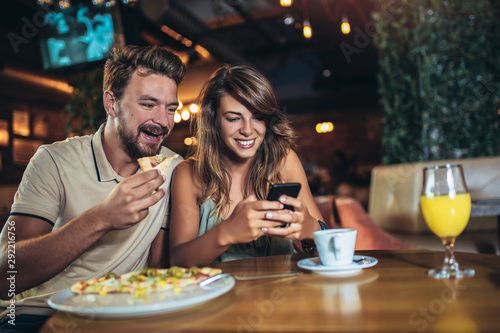 Shot of a young happy couple using phone and eating pizza in a restaurant. Selective focus.