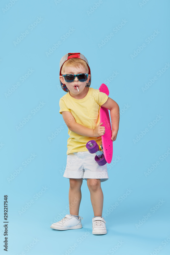 Stylish little toddler child boy in glasses with skateboard having fun over blue background
