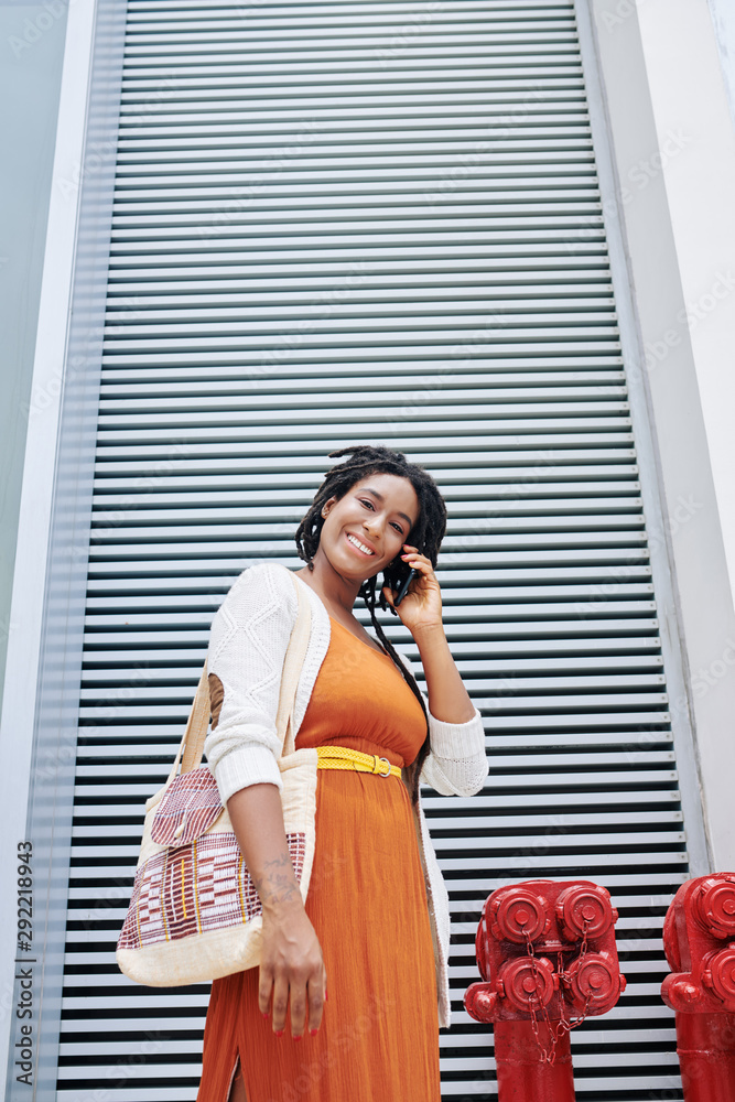 Portrait of African beautiful woman in orange dress talking on mobile phone and smiling at camera while standing in the city