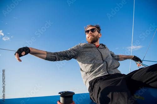 Murais de parede Positive skilled young male sailor with beard wearing sunglasses adjusting sail on boat and examining it while fixing sail on boom