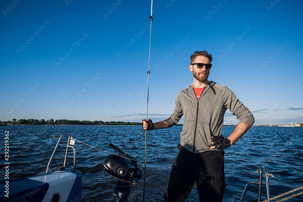 Yacht captain with a beard stands on sail boom on a sailing yacht, holding the rope in his hand and smiling, feeling happy. Adult yachtsman travelling around the world. Copy space Yacht captain with a beard stands on sail boom on a sailing yacht, holding the rope in his hand and smiling, feeling happy. Adult yachtsman travelling around the world. Copy space