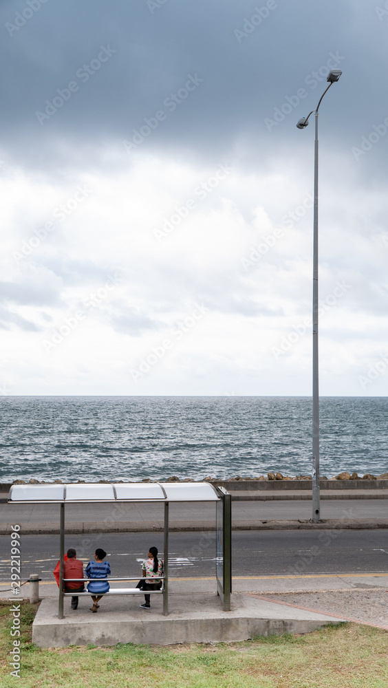 Obraz premium Three people waiting for the bus at a bus stop facing the sea, with a lighting pole