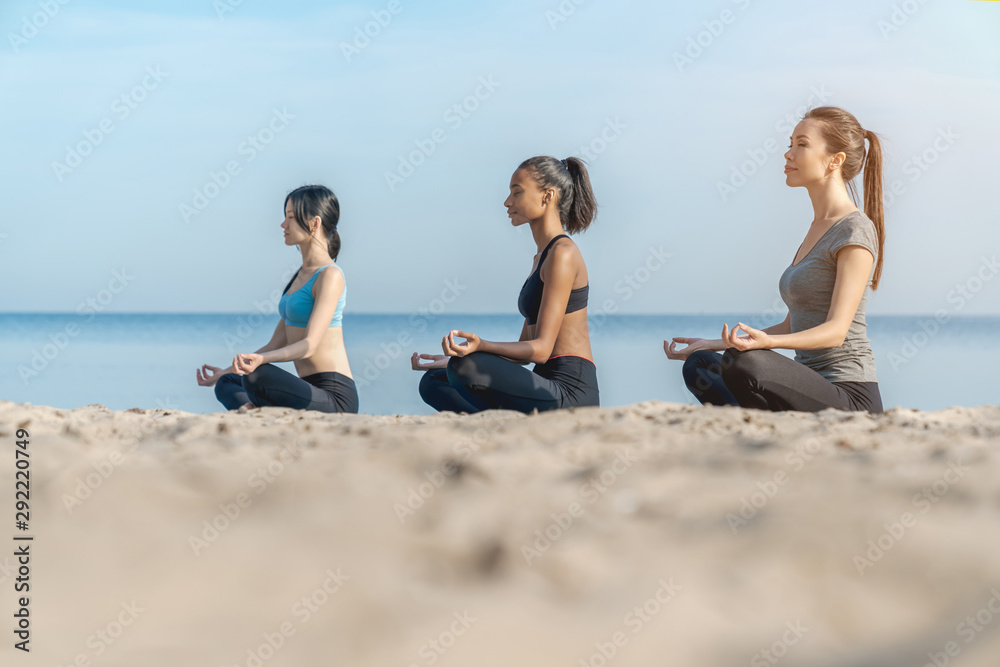 Side view shot of women making yoga meditation in lotus pose on sunny beach near water