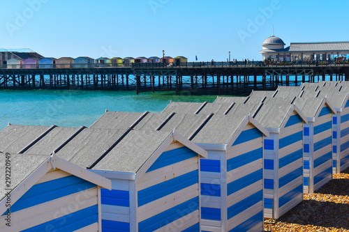 Beach huts on the British coastline on a sunny day. Bright blue and white striped wooden huts an a shingle beach. Pier with colourful beach shacks at the background. Sussex, South East England