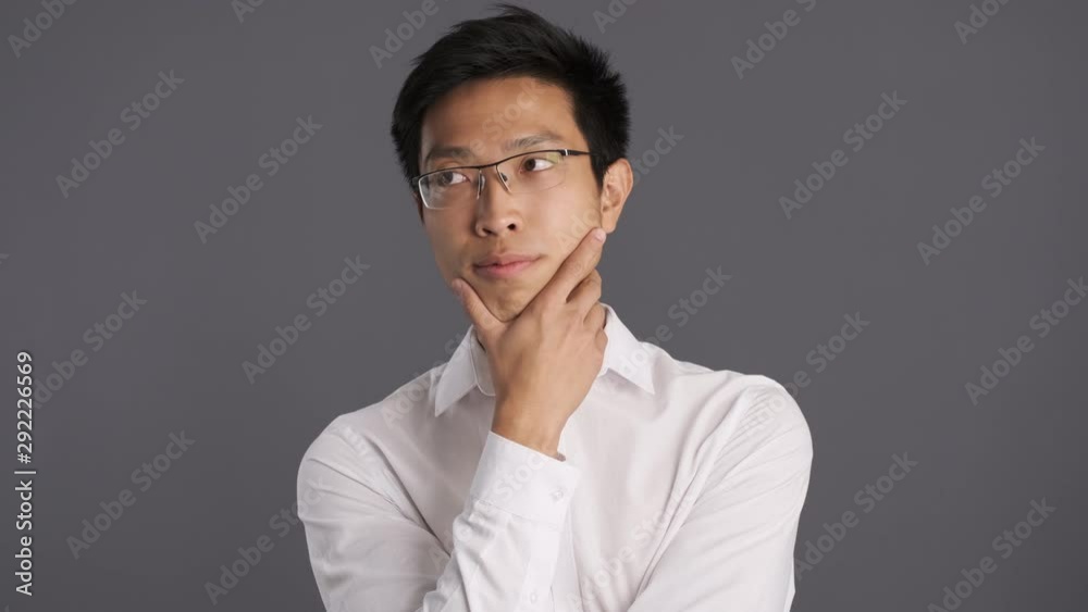 Young pensive asian man in eyeglasses thoughtfully looking in camera over gray background Young pensive asian man in eyeglasses thoughtfully looking in camera over gray background