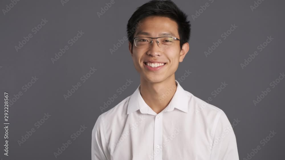 Young smiling asian man in eyeglasses happily looking in camera over gray background Young smiling asian man in eyeglasses happily looking in camera over gray background