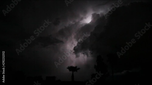 Lightening strikes on a stormy night. Scene goes from pitch black to lit up clouds with distant rain and silhouetted trees and palm trees. Several flashes light up clouds some intense strikes too.