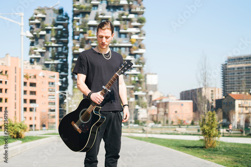 man holding a guitar is standing in front of two green skyscreapers photo