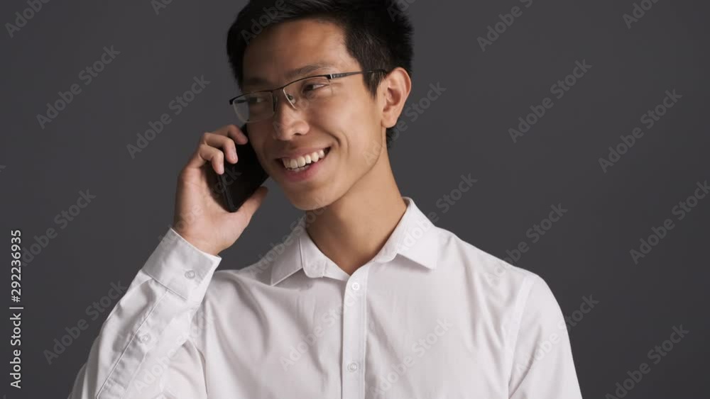 Young attractive smiling asian man in eyeglasses happily talking on cellphone over gray background Young attractive smiling asian man in eyeglasses happily talking on cellphone over gray background