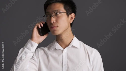 Young serious asian man in eyeglasses tediously talking on cellphone over gray background  photo