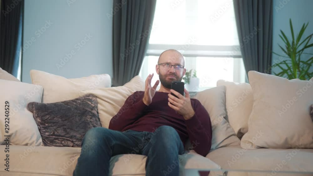 Handsome bearded man in glasses smiling, video call on the phone from his home. He cheerfully says and gestures. He calling to the friends. Stedicam shot. Handsome bearded man in glasses smiling, video call on the phone from his home. He cheerfully says and gestures. He calling to the friends. Stedicam shot.