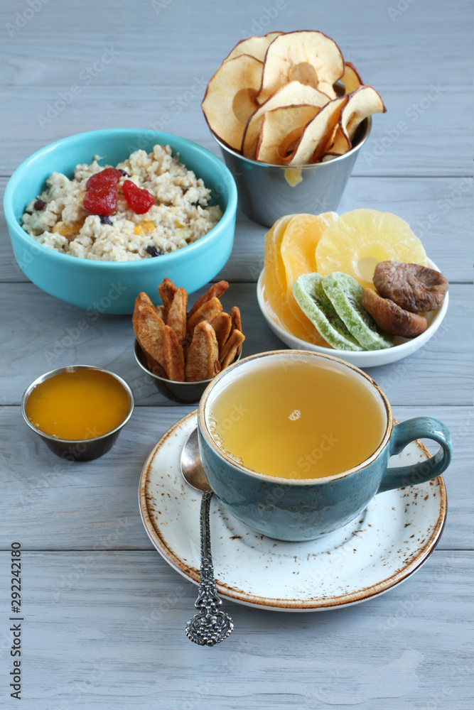 Oatmeal with fruit pieces and candied strawberries, pieces of candied pineapple, kiwi and mango, dried figs, apple and pear chips, honey in small bowls and a cup of green tea on a gray wooden. Closeup Oatmeal with fruit pieces and candied strawberries, pieces of candied pineapple, kiwi and mango, dried figs, apple and pear chips, honey in small bowls and a cup of green tea on a gray wooden. Closeup