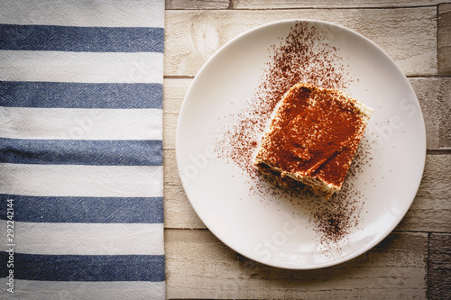 Top view of a slice of homemade tiramisu (traditional Italian dessert) on a white plate on a wooden board with a white and blue kitchen cloth.