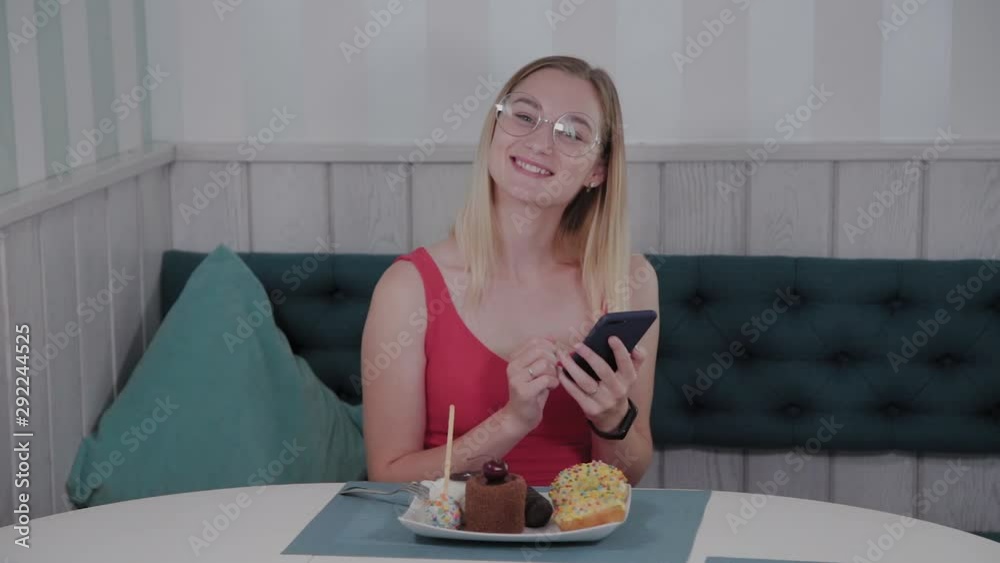 A beautiful young girl uses a mobile phone in a restaurant at a table, around her is a plate with desserts. A beautiful young girl uses a mobile phone in a restaurant at a table, around her is a plate with desserts.