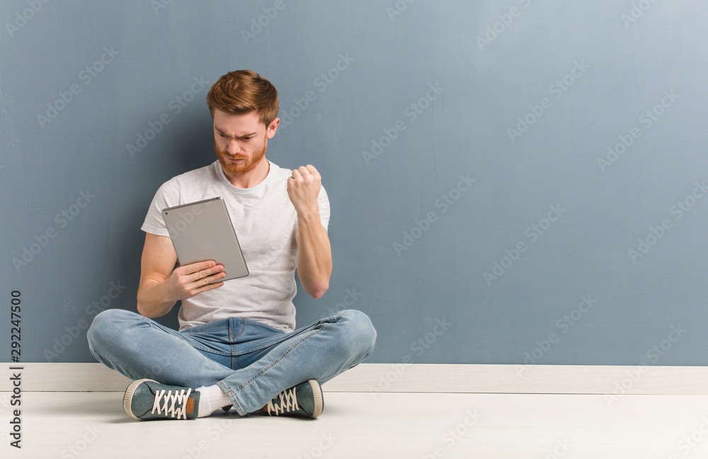 Young redhead student man sitting on the floor showing fist to front, angry expression. He is holding a tablet. Young redhead student man sitting on the floor showing fist to front, angry expression. He is holding a tablet.