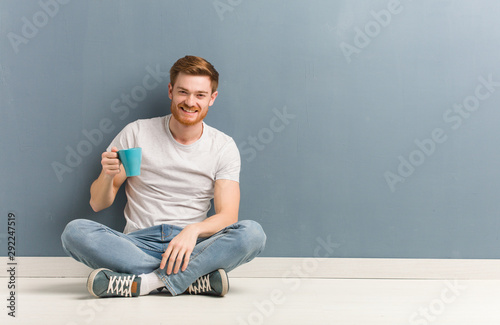 Young redhead student man sitting on the floor cheerful with a big smile. He is holding a coffee mug.