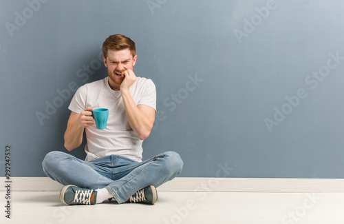 Young redhead student man sitting on the floor biting nails, nervous and very anxious. He is holding a coffee mug.