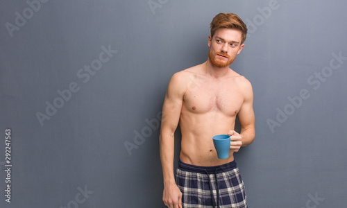 Young shirtless redhead man smiling confident and crossing arms, looking up. He is holding a coffee mug.