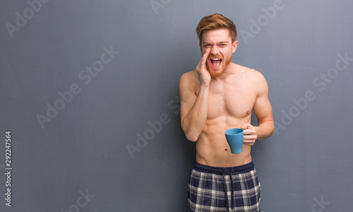 Young shirtless redhead man shouting something happy to the front. He is holding a coffee mug.