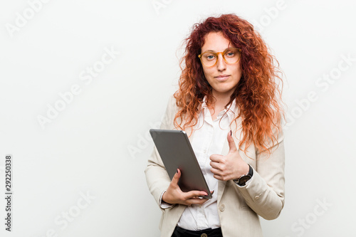 Young caucasian business redhead woman holding a tablet smiling and raising thumb up