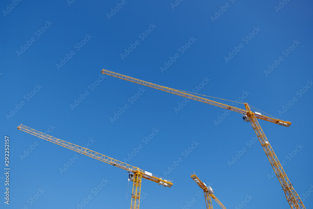 Low angle view group of three yellow tower cranes work over building construction site against clear blue sky. Low angle view group of three yellow tower cranes work over building construction site against clear blue sky.