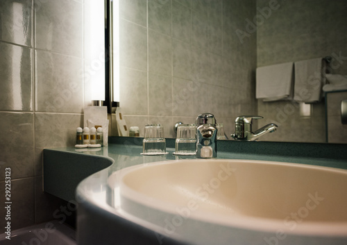 Interior of modern three star hotel with sink  chrome water tap  two glasses and defocused shampoo in the corner