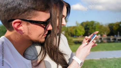 Young attractive couple standing in the park. Using modern mobile phone. Girl showing something on his smartphone. Outdoor. Close up shot.
