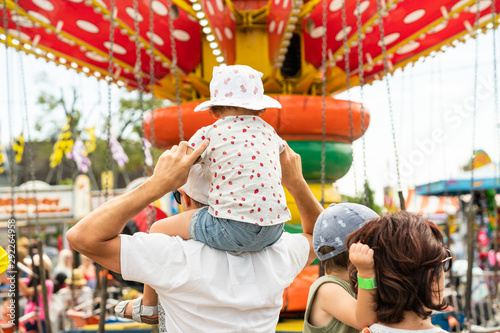 Child sitting on her father's shoulders in front of a colorful carousel at a carnival. Girl and boy with their parents at the vintage amusement park rides. Wearing a cute ladybug outfit and a sun hat.