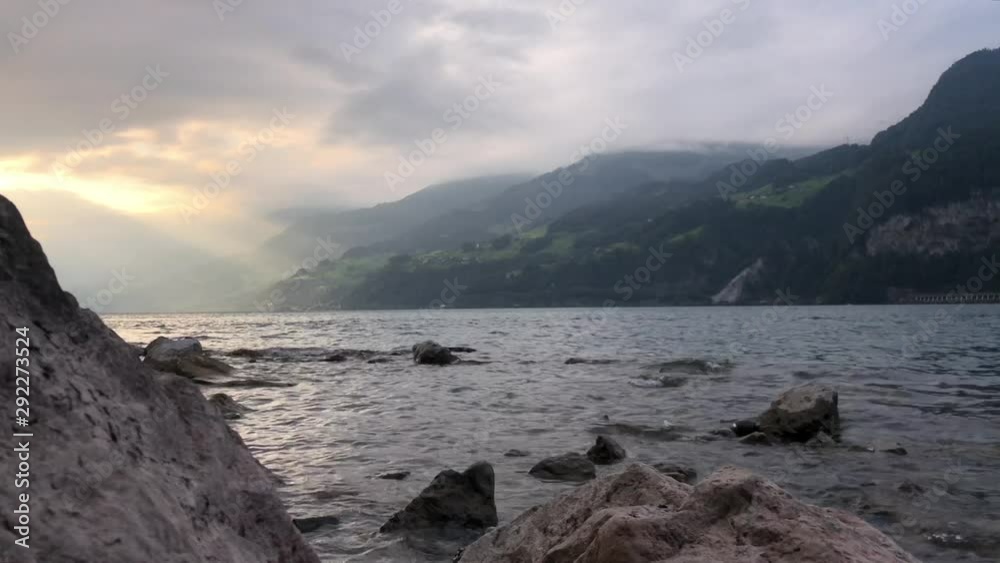 Golden sun rays peeking through the clouds and shining onto the mountainside as the water laps over the rocks. Golden sun rays peeking through the clouds and shining onto the mountainside as the water laps over the rocks.