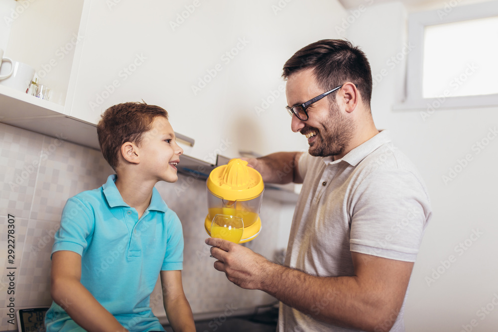 Happy father and kid standing in kitchen in embrace. They holding squeezed juice in hands Happy father and kid standing in kitchen in embrace. They holding squeezed juice in hands