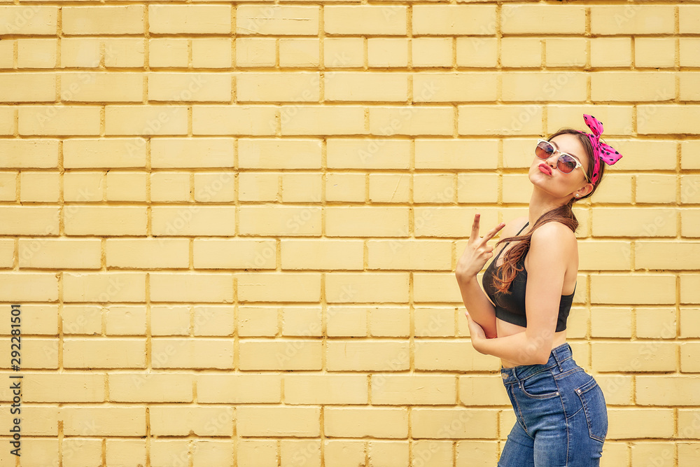 cheerful girl with a bow and a black tank top emotionally posing in sunglasses and jeans on a background of a yellow brick wall. copy space.