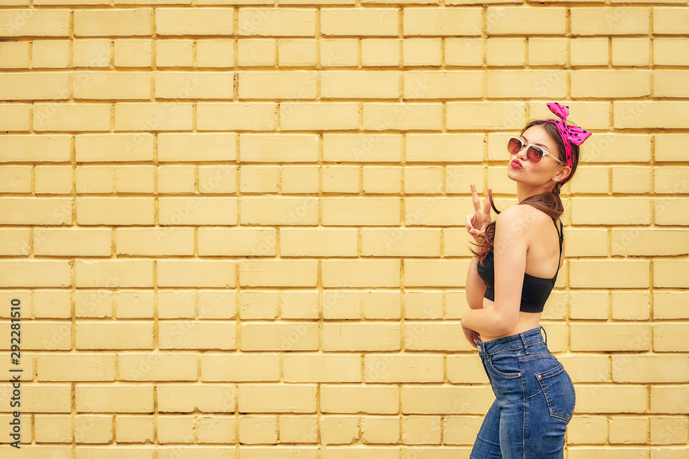 cheerful girl with a bow and a black tank top emotionally posing in sunglasses and jeans on a background of a yellow brick wall. copy space.