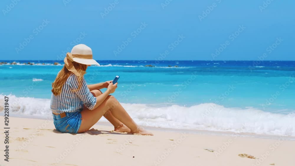Young woman using phone sitting on sandy Caribbean beach with waves Young woman using phone sitting on sandy Caribbean beach with waves