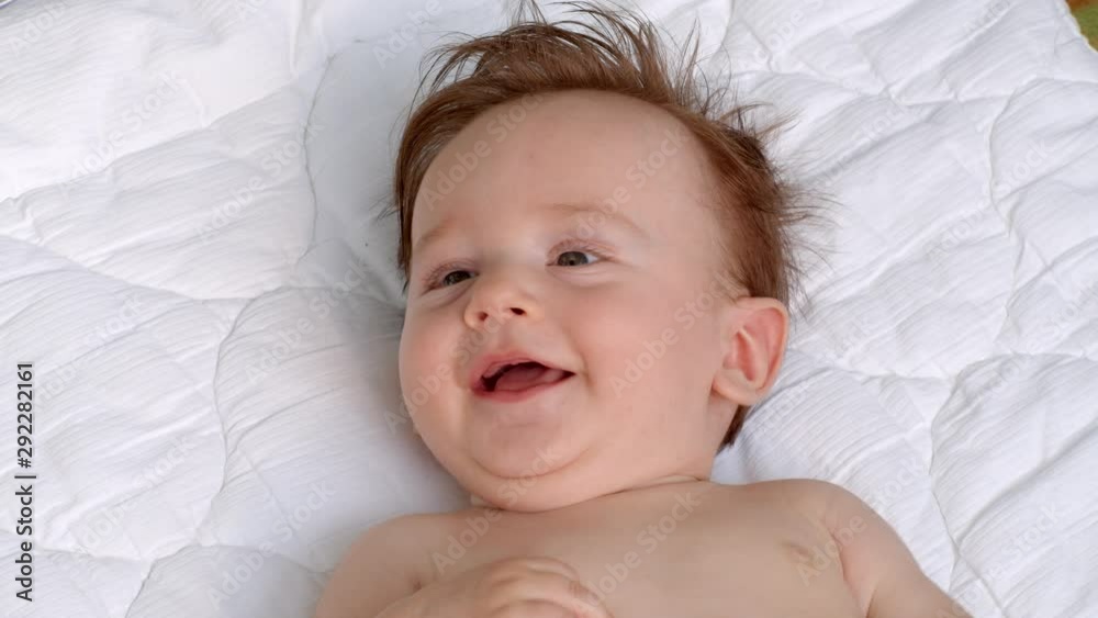 White smiling little child with dark thick hair and dark eyes is lying topless on the blanket on open air and observes the area. It is moving head and fingers. It raises left and right hand White smiling little child with dark thick hair and dark eyes is lying topless on the blanket on open air and observes the area. It is moving head and fingers. It raises left and right hand