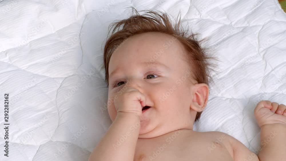 White little smiling child with dark thick hair and dark eyes is lying topless on the blanket on open air and sucks thumb of right hand moving its head and fingers. It raises left hand White little smiling child with dark thick hair and dark eyes is lying topless on the blanket on open air and sucks thumb of right hand moving its head and fingers. It raises left hand
