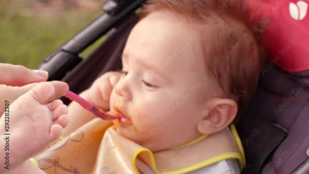 White excited infant with dark hair and dirty face is sitting in stroller. Mother feeds it by spoon. It looks to be surprised and holds its foot high White excited infant with dark hair and dirty face is sitting in stroller. Mother feeds it by spoon. It looks to be surprised and holds its foot high