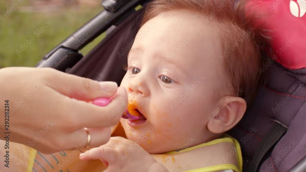 White excited infant with dark hair and dirty face is sitting in stroller. Mother feeds it by spoon. It looks to be surprised and holds its foot high smiling White excited infant with dark hair and dirty face is sitting in stroller. Mother feeds it by spoon. It looks to be surprised and holds its foot high smiling